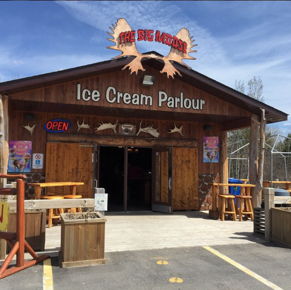 The log-cabin storefront of Big Moose Ice Cream Parlour on Great Northern Road, with its antlered wooden sign above the Ice Cream Parlour entrance and covered deck with wooden stools