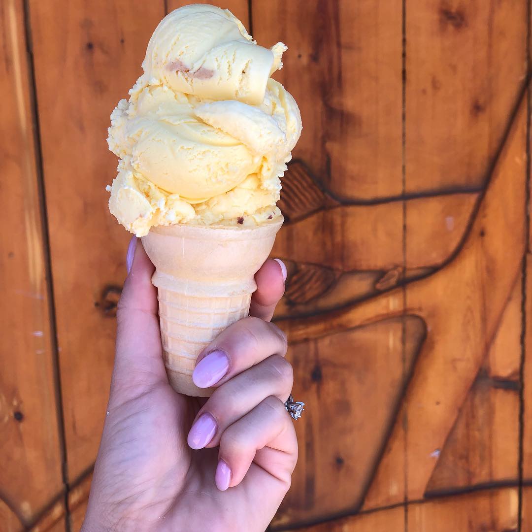A double scoop of creamy pale-yellow frozen yogurt in a sugar cone, held against the wooden log-cabin wall
